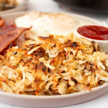Crispy hash browns on a plate with bacon, eggs, and ketchup.