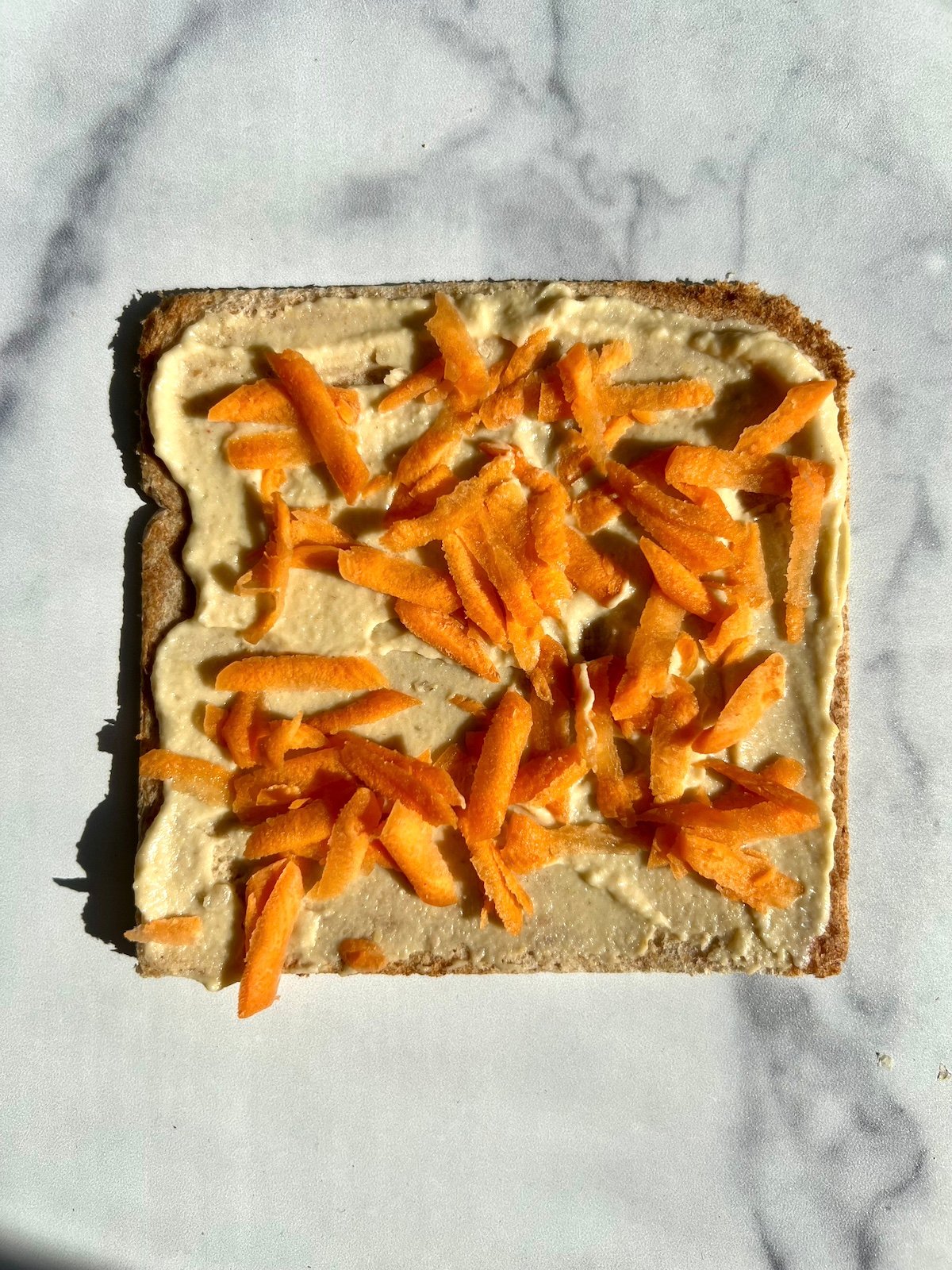 Piece of sandwich bread with hummus and grated carrots on top.