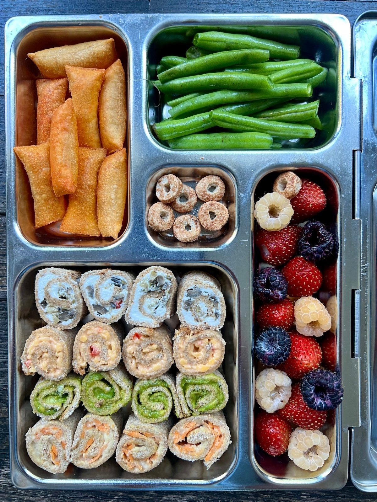 Lunch box filled with sushi sandwiches, berries, green beans, chips and sweet treat.