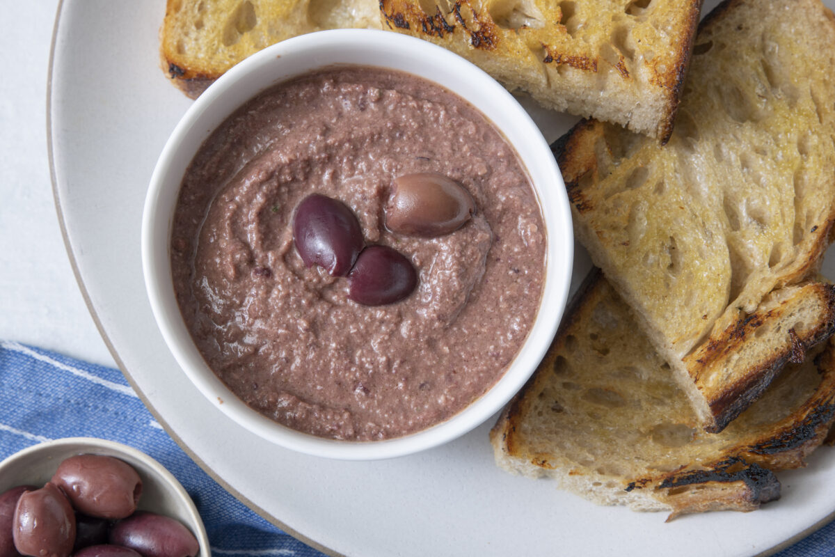 Olive tapenade in small bowl on serving platter with crispy bread.