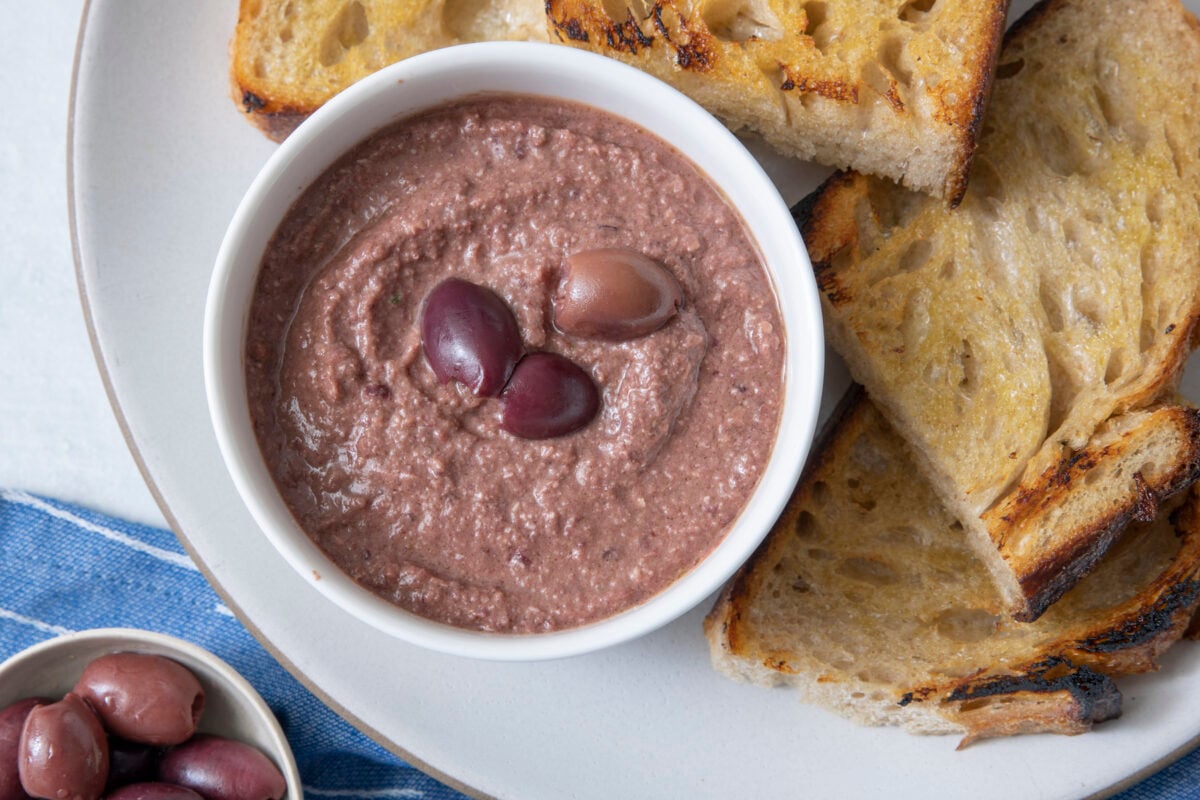 Olive tapenade in small bowl on serving platter with crispy bread.