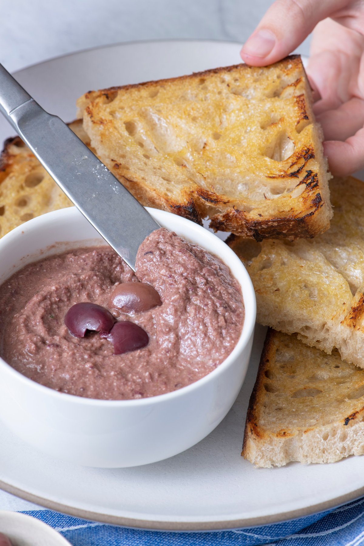 Knife scooping olive tapenade out of small bowl.