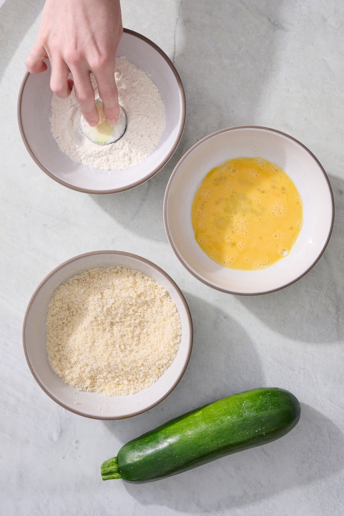 Three bowls. One with flour, another with beaten egg and the third with panko breading. A hand is dipping a zucchini coin in the flour bowl.