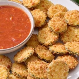 Baked zucchini coins on serving platter with marinara sauce in a bowl on the side.