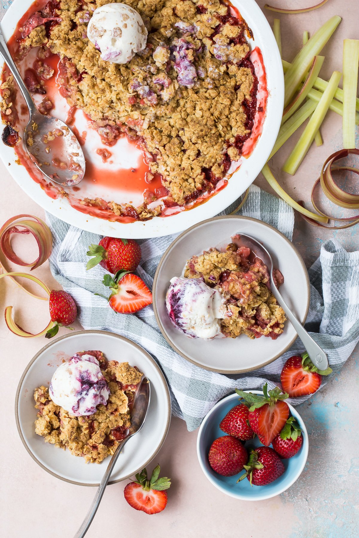 Strawberry rhubarb crumble in pie dish with two servings in bowls topped with ice cream.