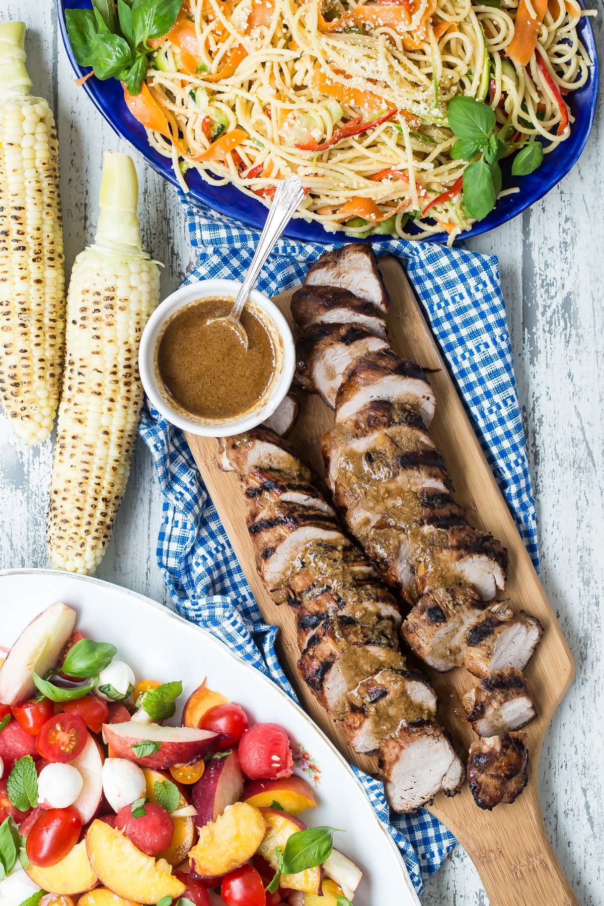 southern style pork tenderloin on cutting board surrounded by sides of grilled corn, pasta and summer salad