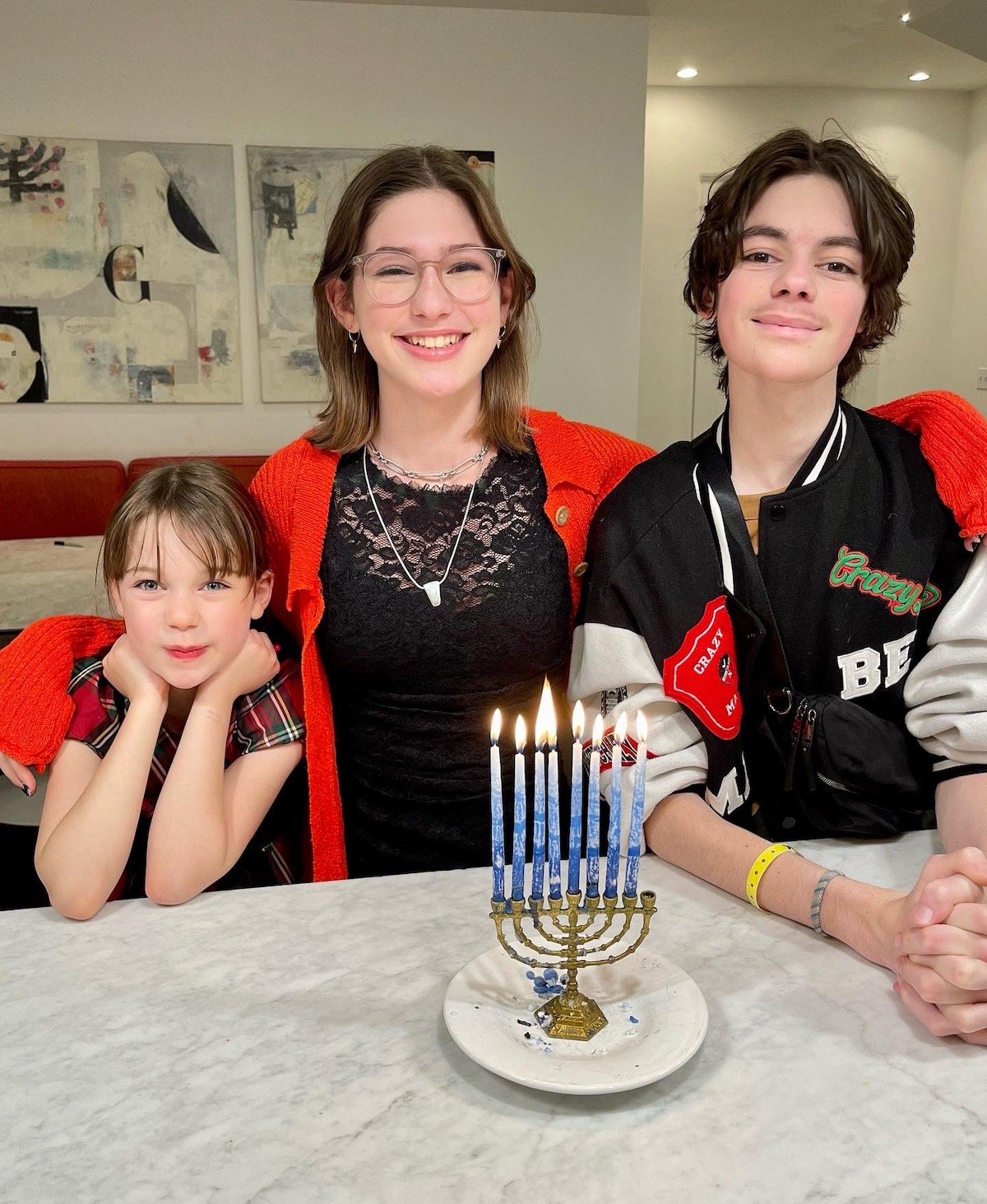 Three siblings smiling for the camera lighting a menorah. 