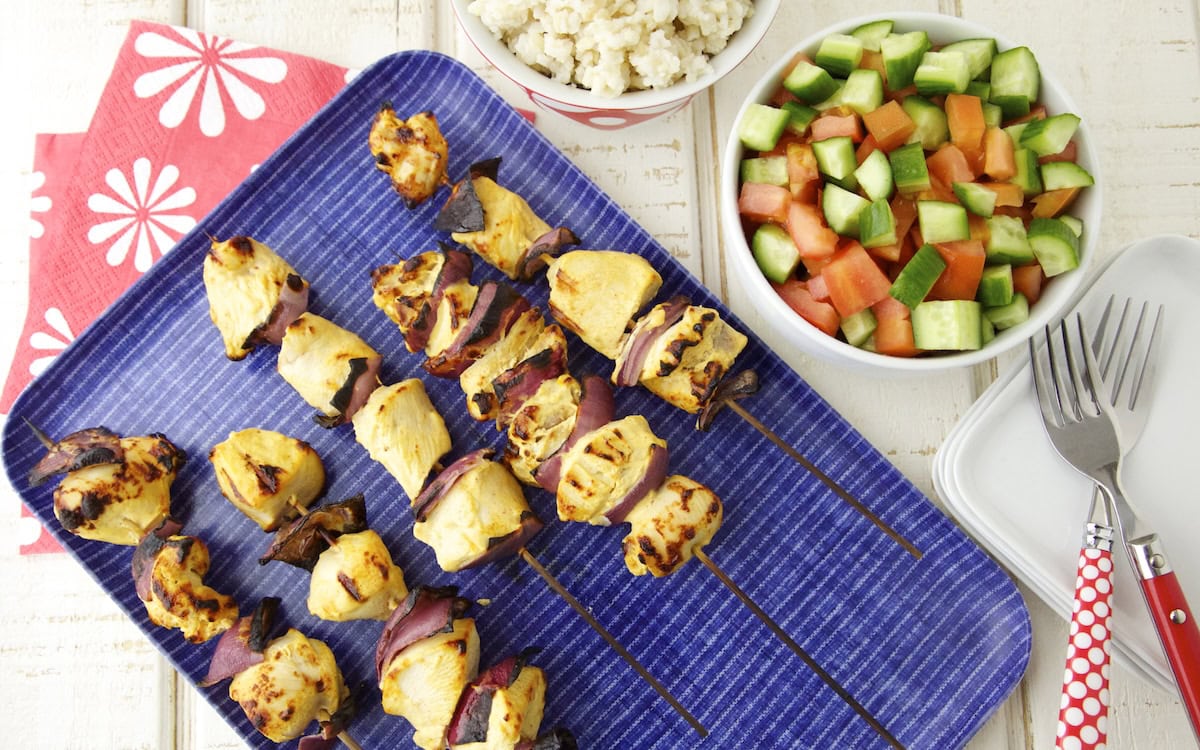 Grilled chicken and vegetable skewers on a blue tray, with bowls of salad and mashed potatoes, plus red-handled forks and red napkins with white floral designs on a white wooden table.