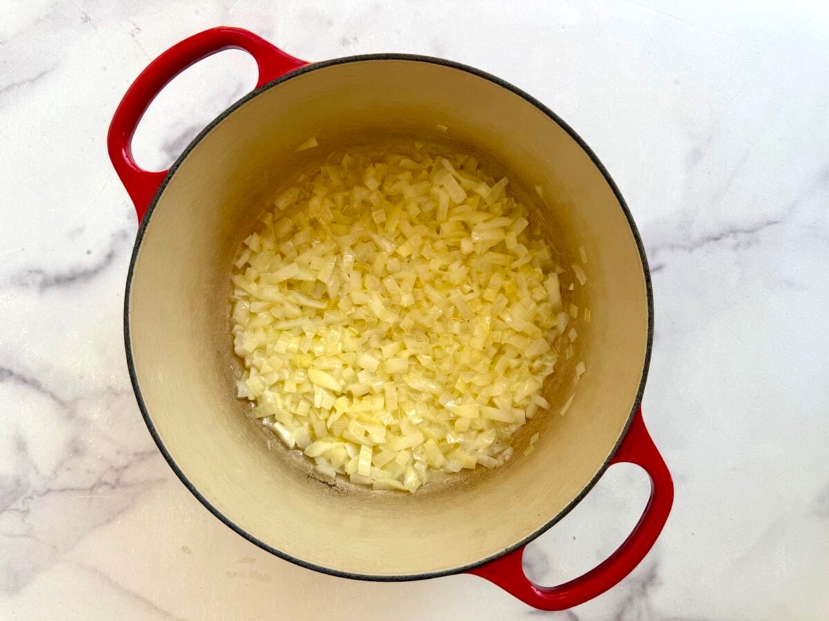 Diced onions cooking in a red enameled cast iron Dutch oven on a white marble surface.