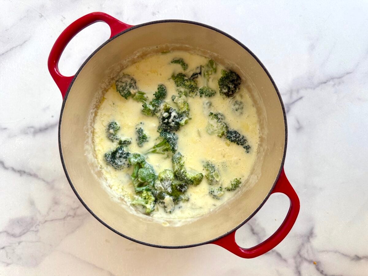 Broccoli florets simmering in a creamy sauce inside a red enameled cast iron pot.