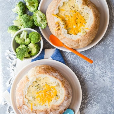 Two bread bowls filled with creamy broccoli cheese soup, served with side bowls of fresh broccoli florets and spoons.