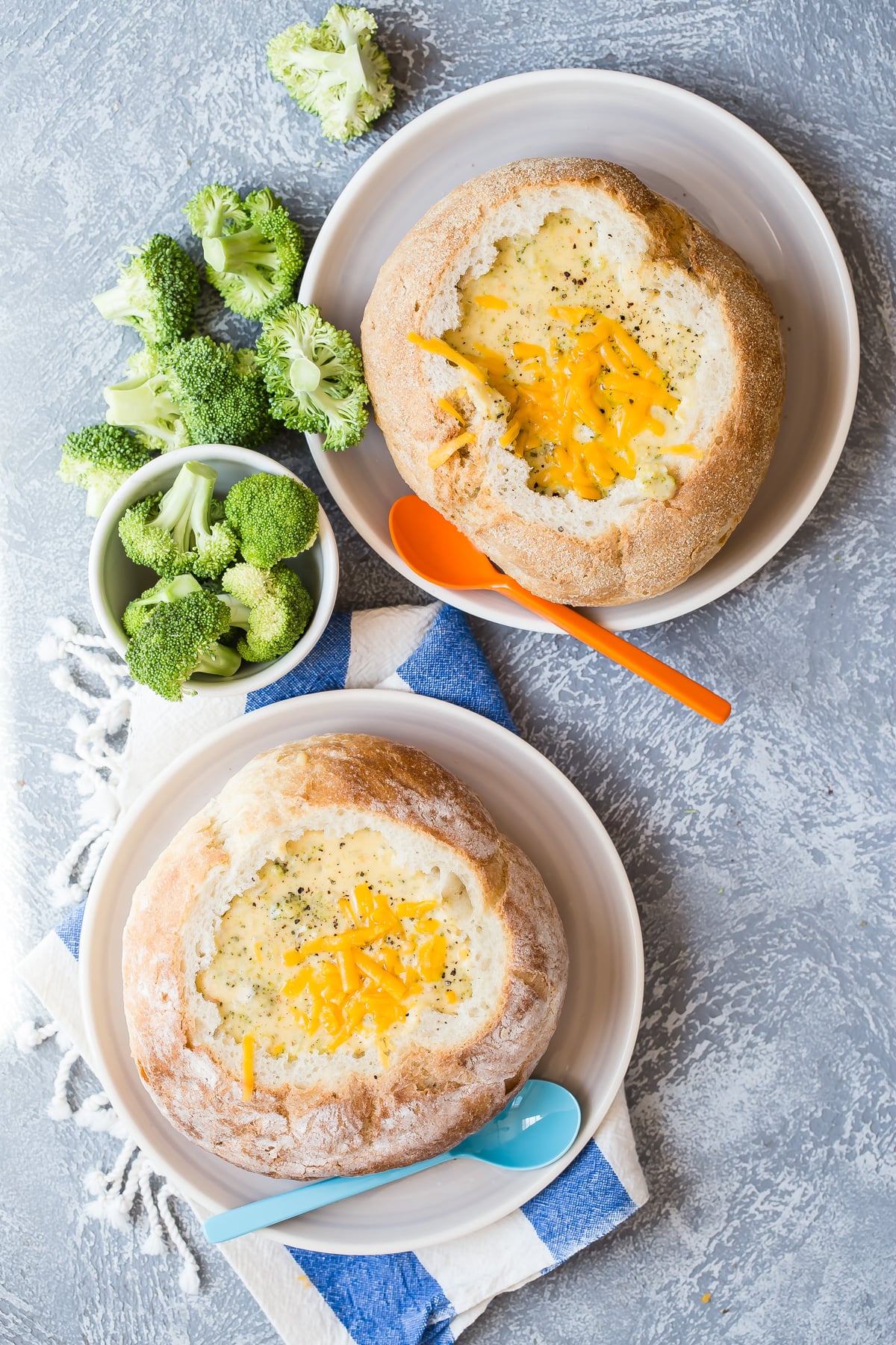Two bread bowls filled with creamy broccoli soup, served with side bowls of fresh broccoli florets and spoons.