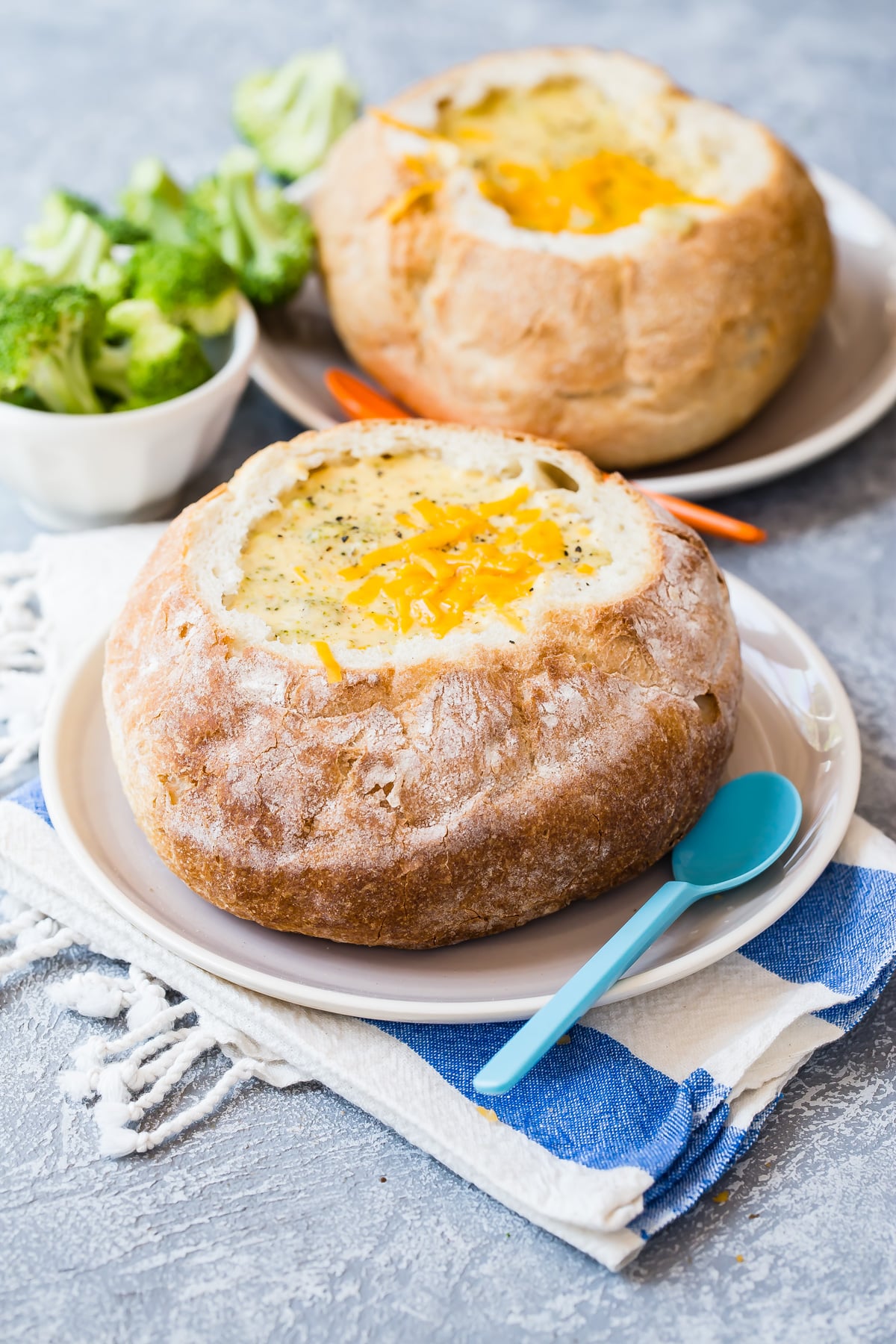 Broccoli cheddar soup served in a hollowed-out bread bowl with a blue spoon, accompanied by a small bowl of broccoli florets.