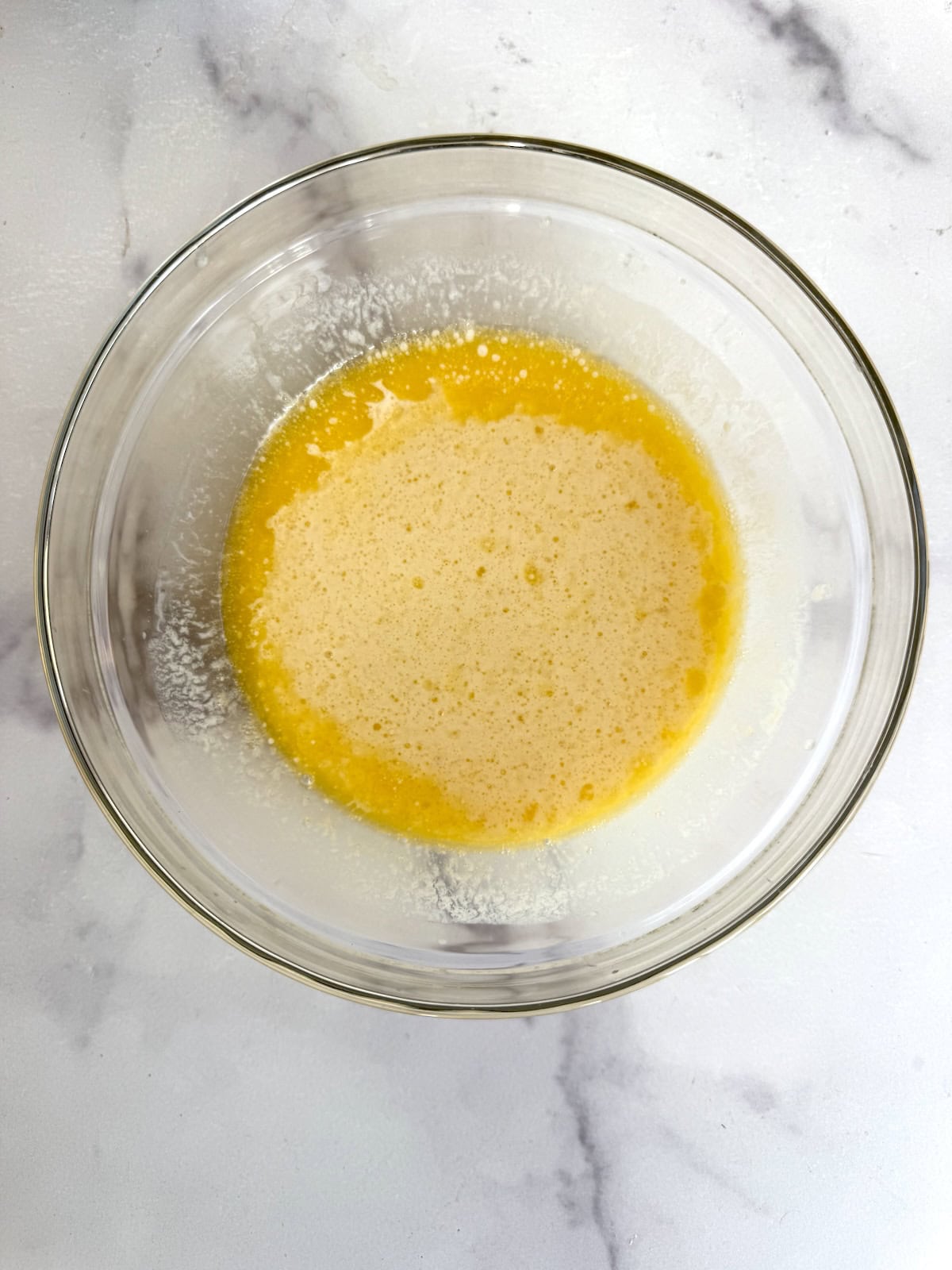 A glass bowl filled with a yellow, frothy liquid mixture sits on a white marble surface.
