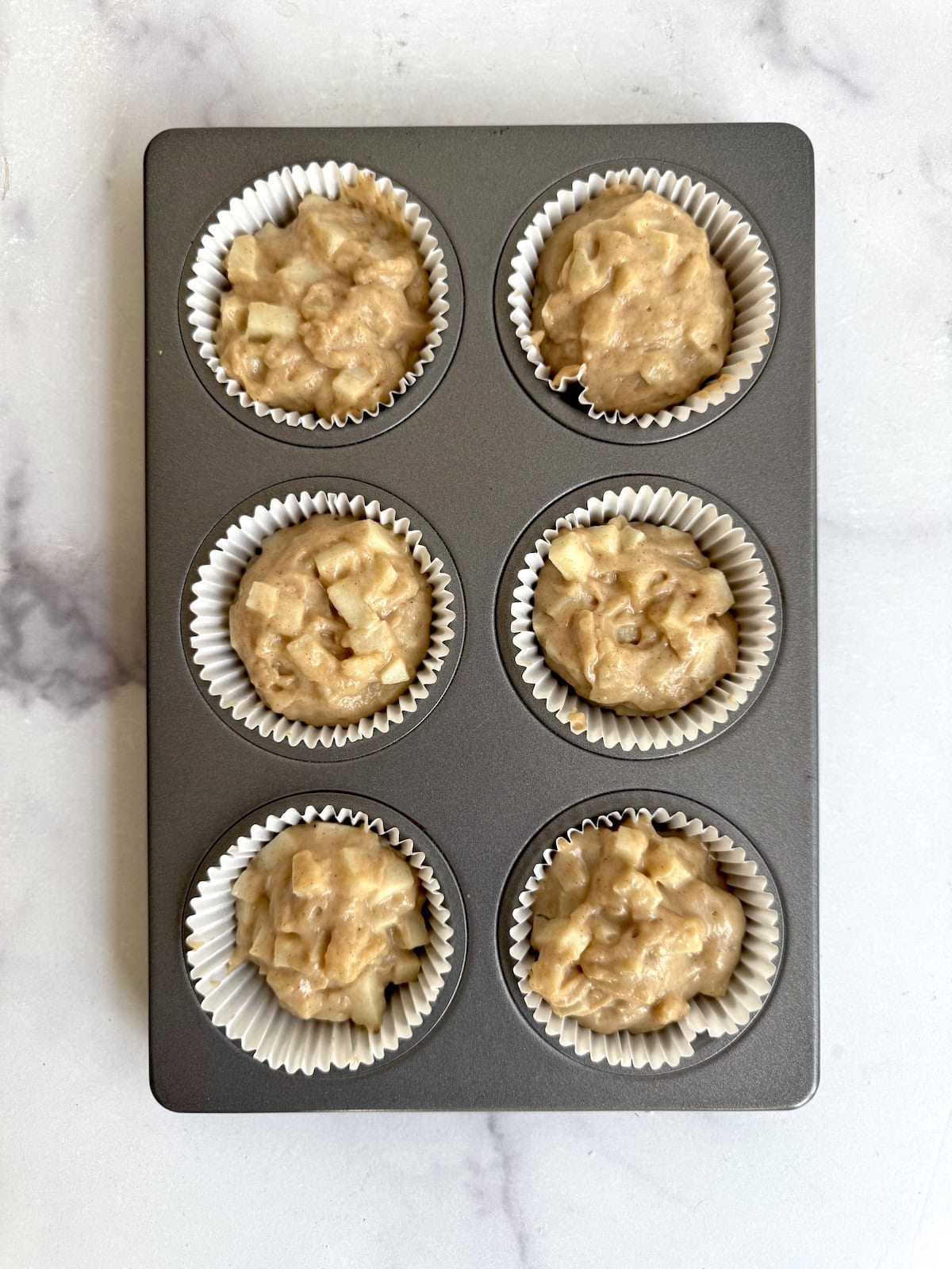 A muffin tin with six paper liners, each filled with unbaked muffin batter containing visible chunks of apple, sits on a white marble surface.
