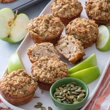A plate of apple muffins with crumb topping, some halved to show the inside, sits beside green apple slices and a small bowl of pumpkin seeds. A muffin tray and apple are in the background.