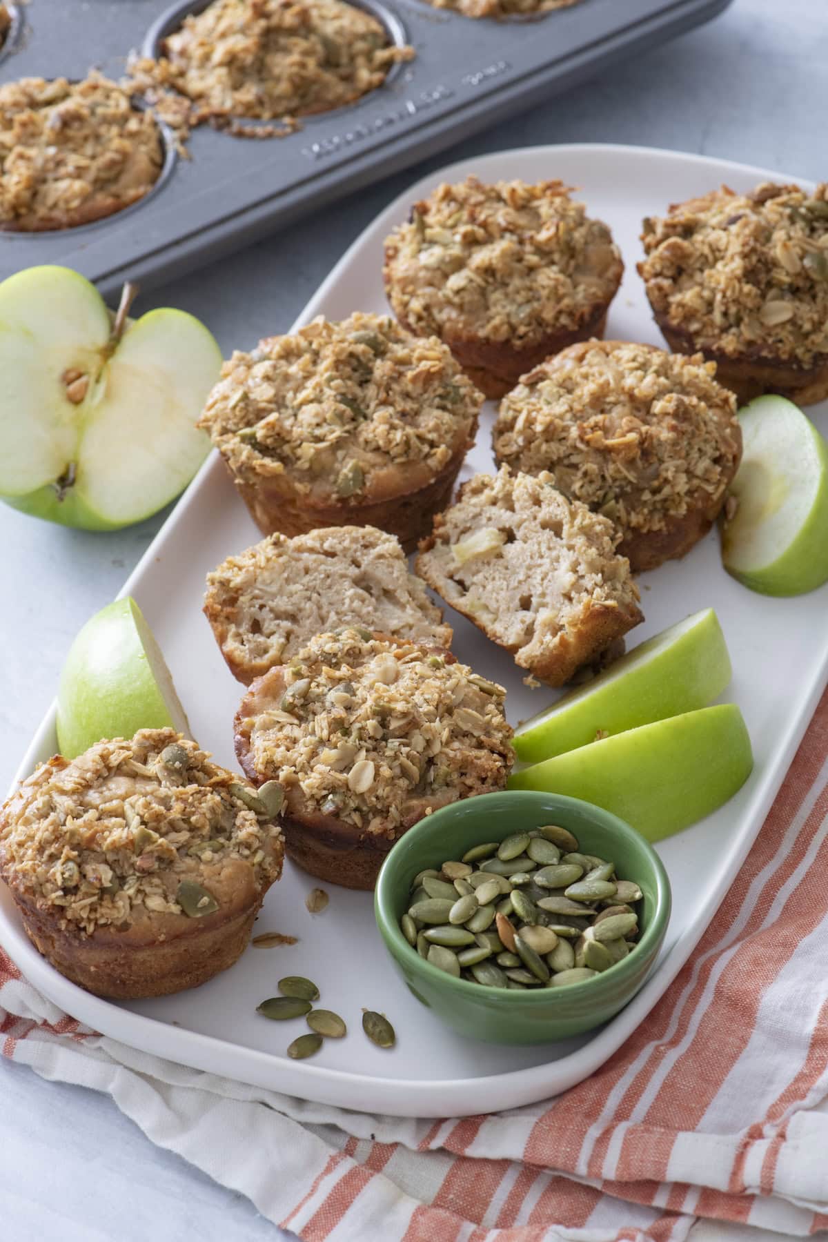 A plate of apple pie muffins with crumb topping, some halved to show the inside, sits beside green apple slices and a small bowl of pumpkin seeds. A muffin tray and apple are in the background.