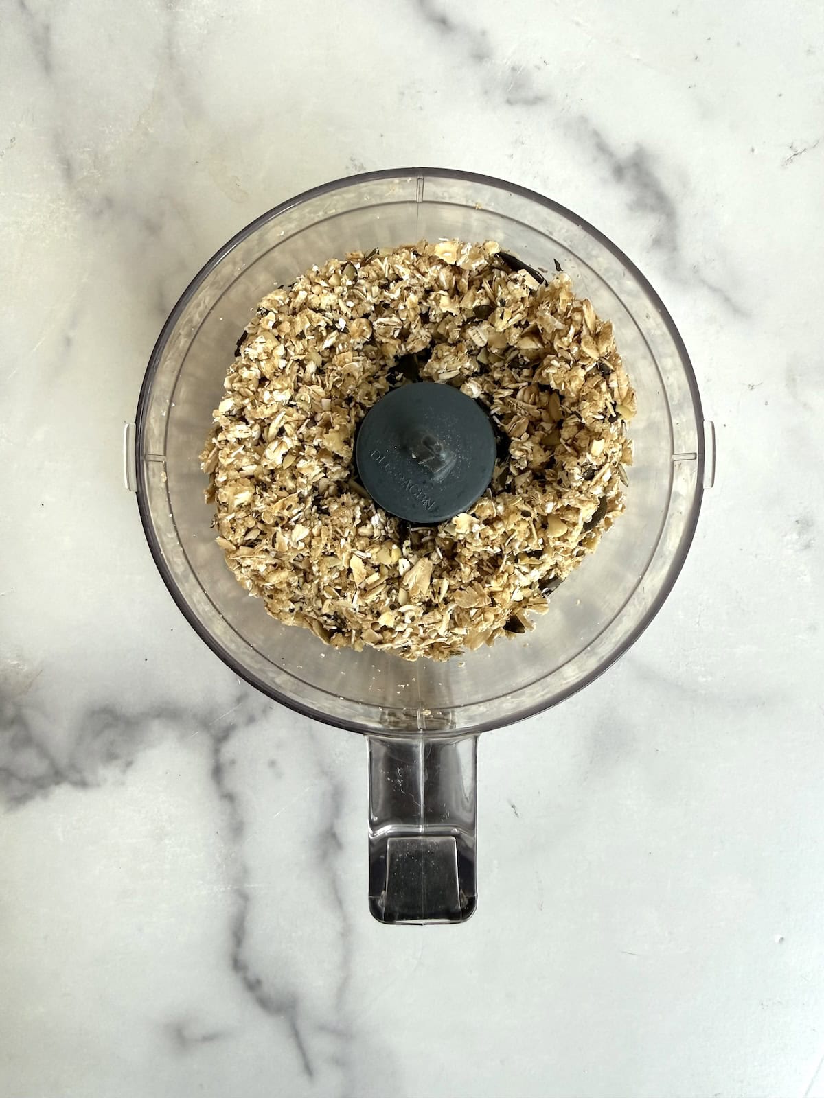 Top view of a clear food processor bowl filled with dry rolled oats, placed on a white marble surface.