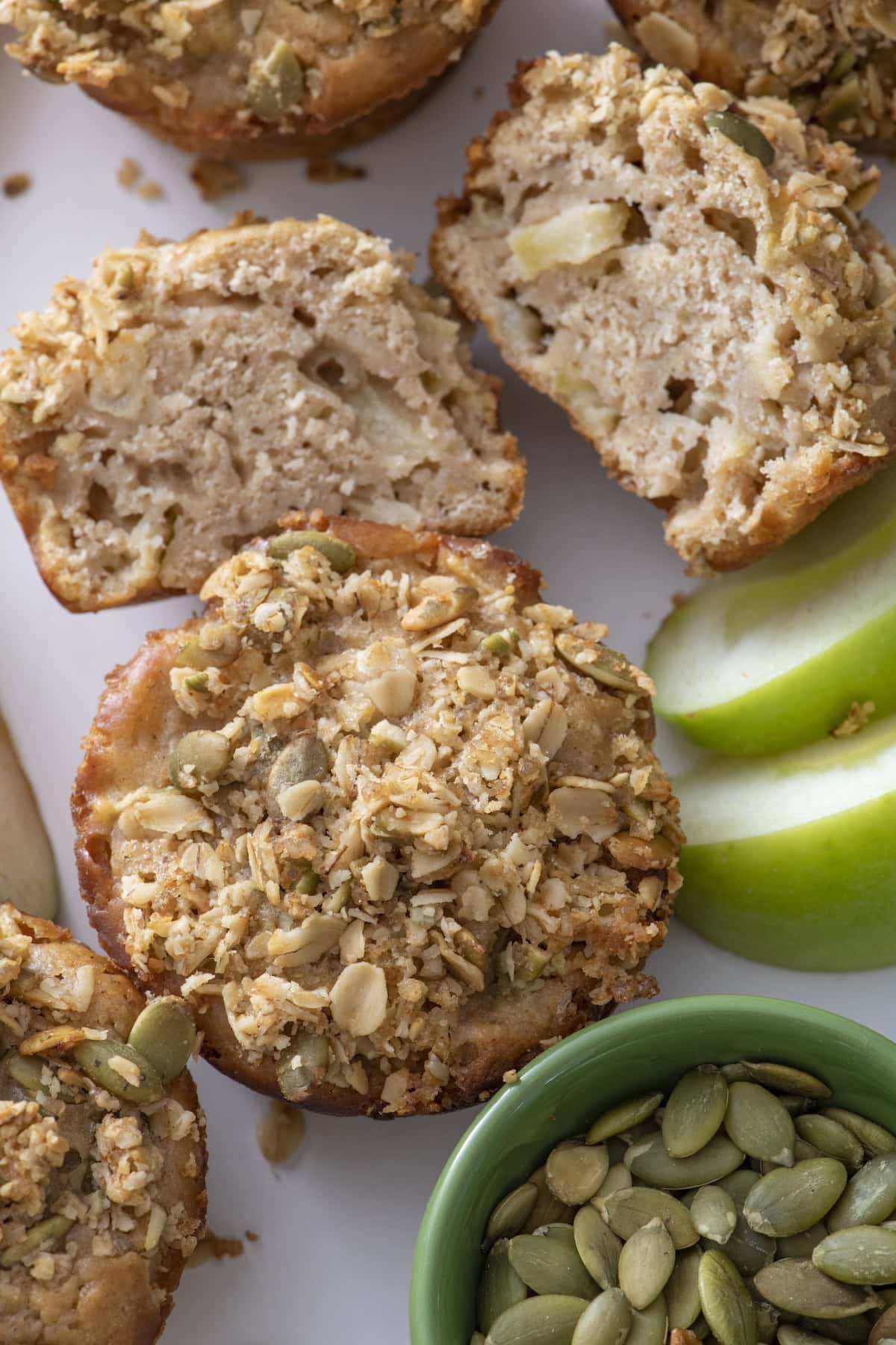 Close-up of apple muffins topped with oats and pumpkin seeds, some cut open to show the moist interior. Green apple slices and a small bowl of pumpkin seeds are also visible on the white surface.