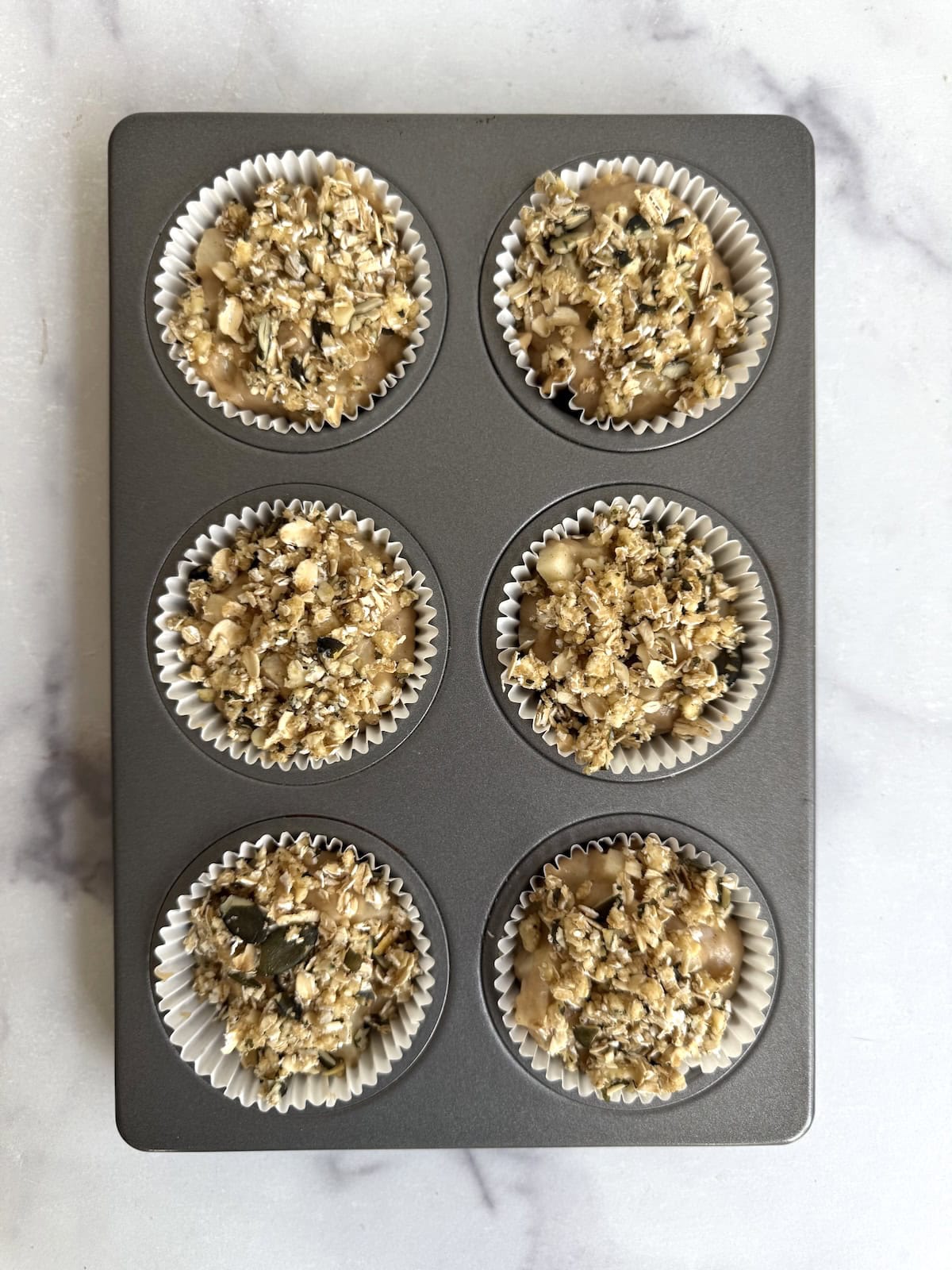 A muffin tin with six paper liners, each filled with unbaked oat and seed mixture, sits on a light marble surface.