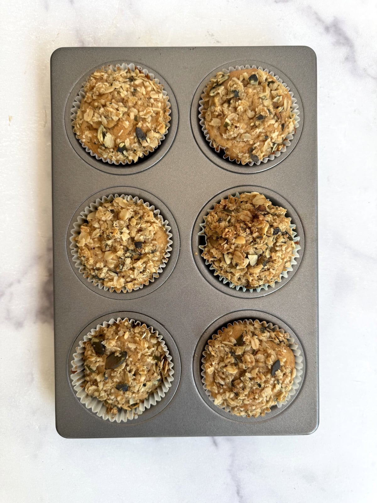 A muffin tin with six paper-lined cups, each filled with baked apple pie muffins topped with seeds and oats, on a light marble surface.