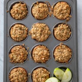 A muffin tin baked apple pie muffins, each topped with oat crumble, sits on a grey surface. In the bottom right corner of the tin are three green apple slices.