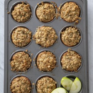 A muffin tin baked apple pie muffins, each topped with oat crumble, sits on a grey surface. In the bottom right corner of the tin are three green apple slices.