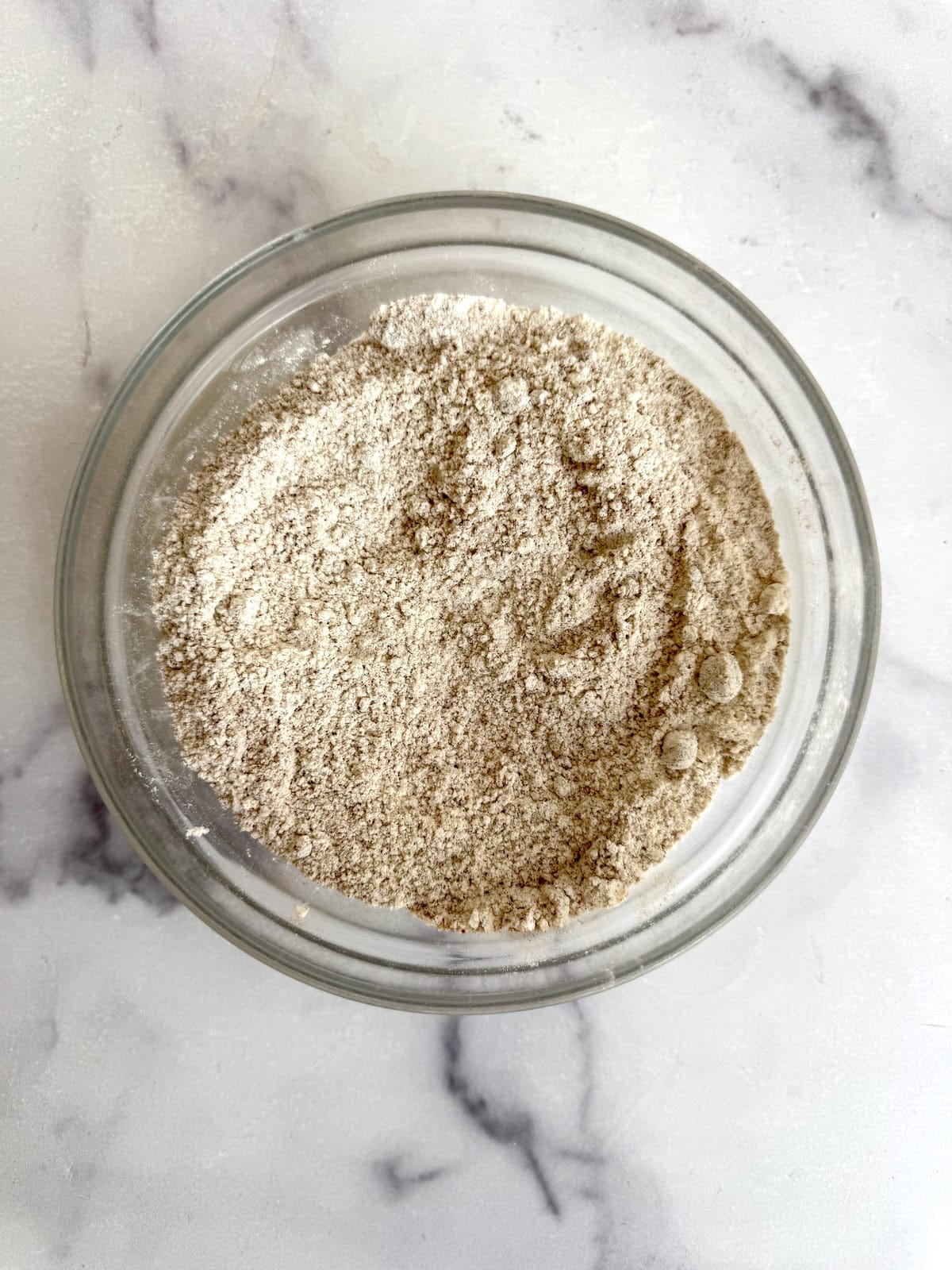 A clear glass bowl filled with a light brown, finely ground powder sits on a white marble surface.