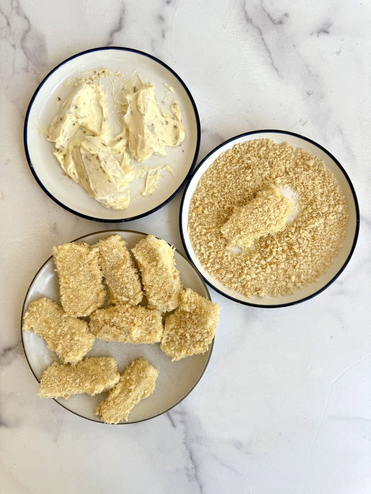 Three plates on a white surface: one with fish pieces in a creamy mixture, one with breadcrumbs and a coated piece, and one with several fully breaded fish strips ready to cook.