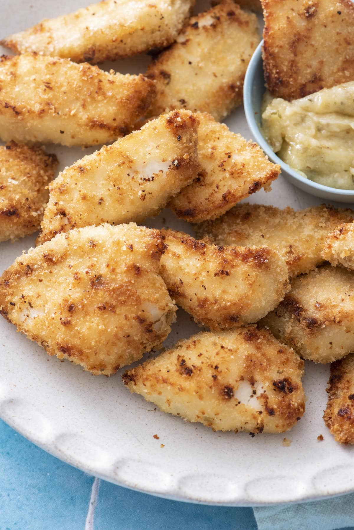 Golden, crispy breaded fish fingers on a white plate, served with a small bowl of light-colored dipping sauce.
