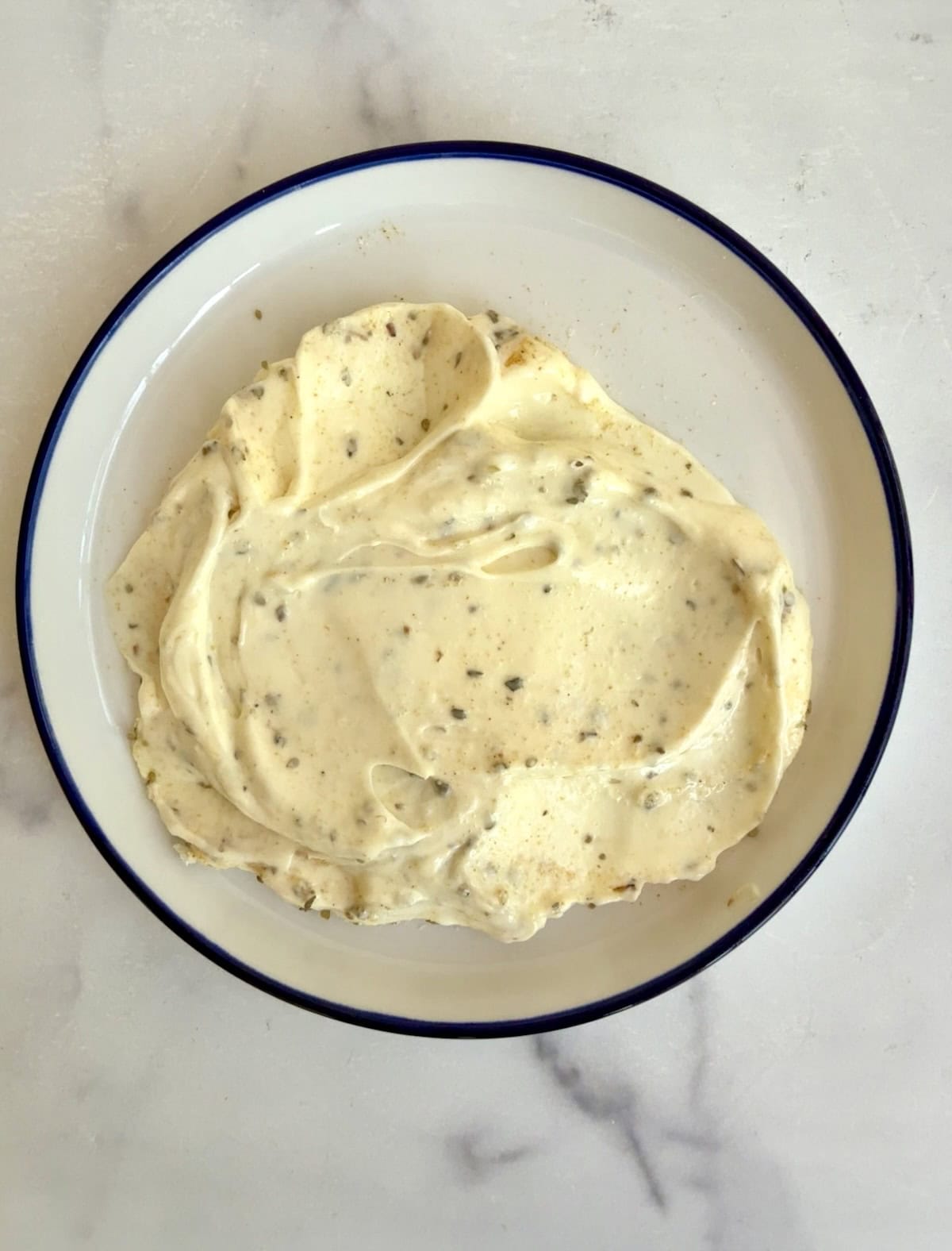 A white plate with a blue rim holds a spread of herb-flecked mayonnaise on a light marble surface.