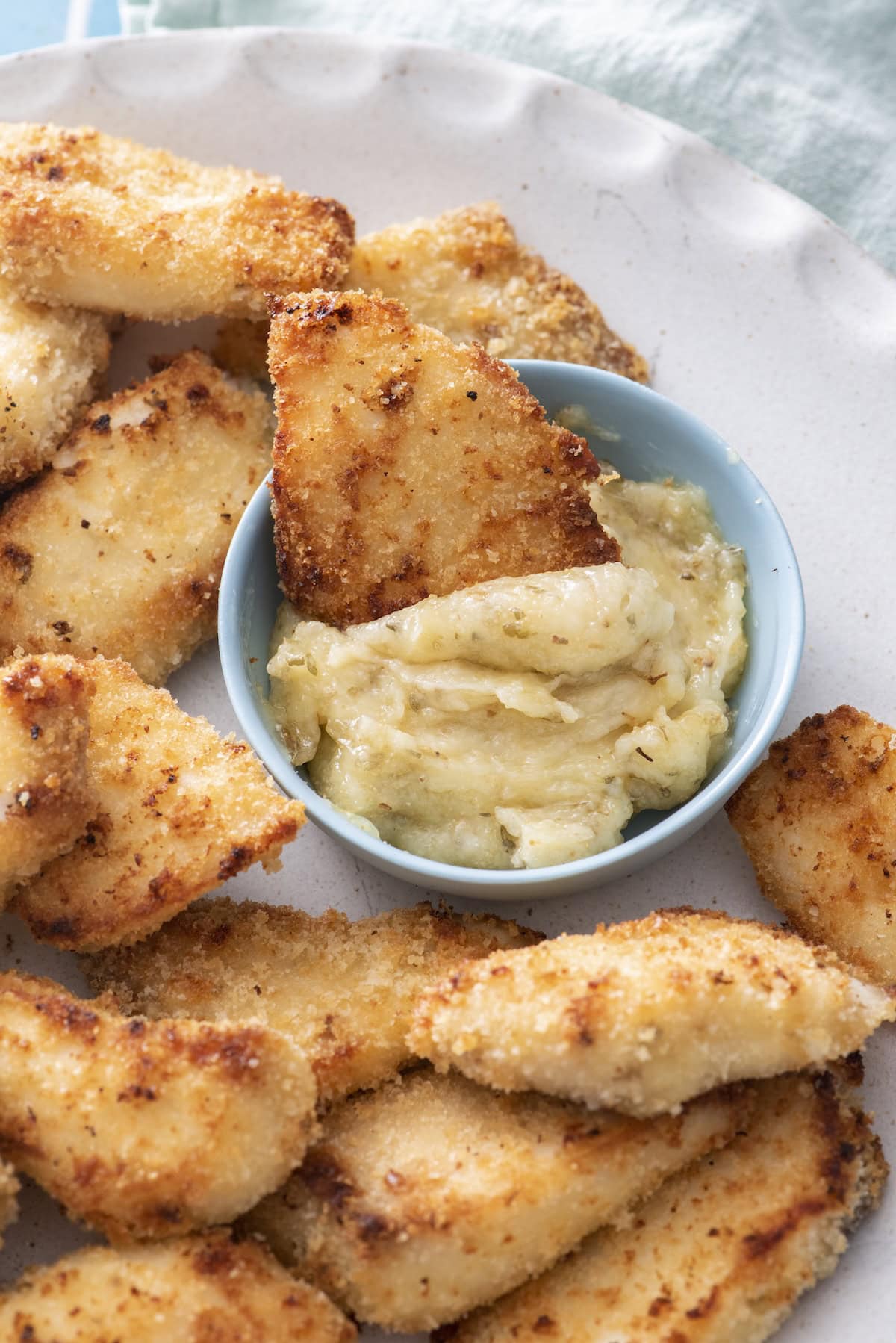 A plate of breaded, golden-brown fish fingers with one piece being dipped into a creamy dip in a small blue bowl.