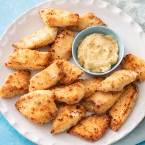 A plate of golden-brown breaded fish pieces with a small bowl of creamy dipping sauce, set on a light blue tiled surface with a pale green cloth napkin under the plate.