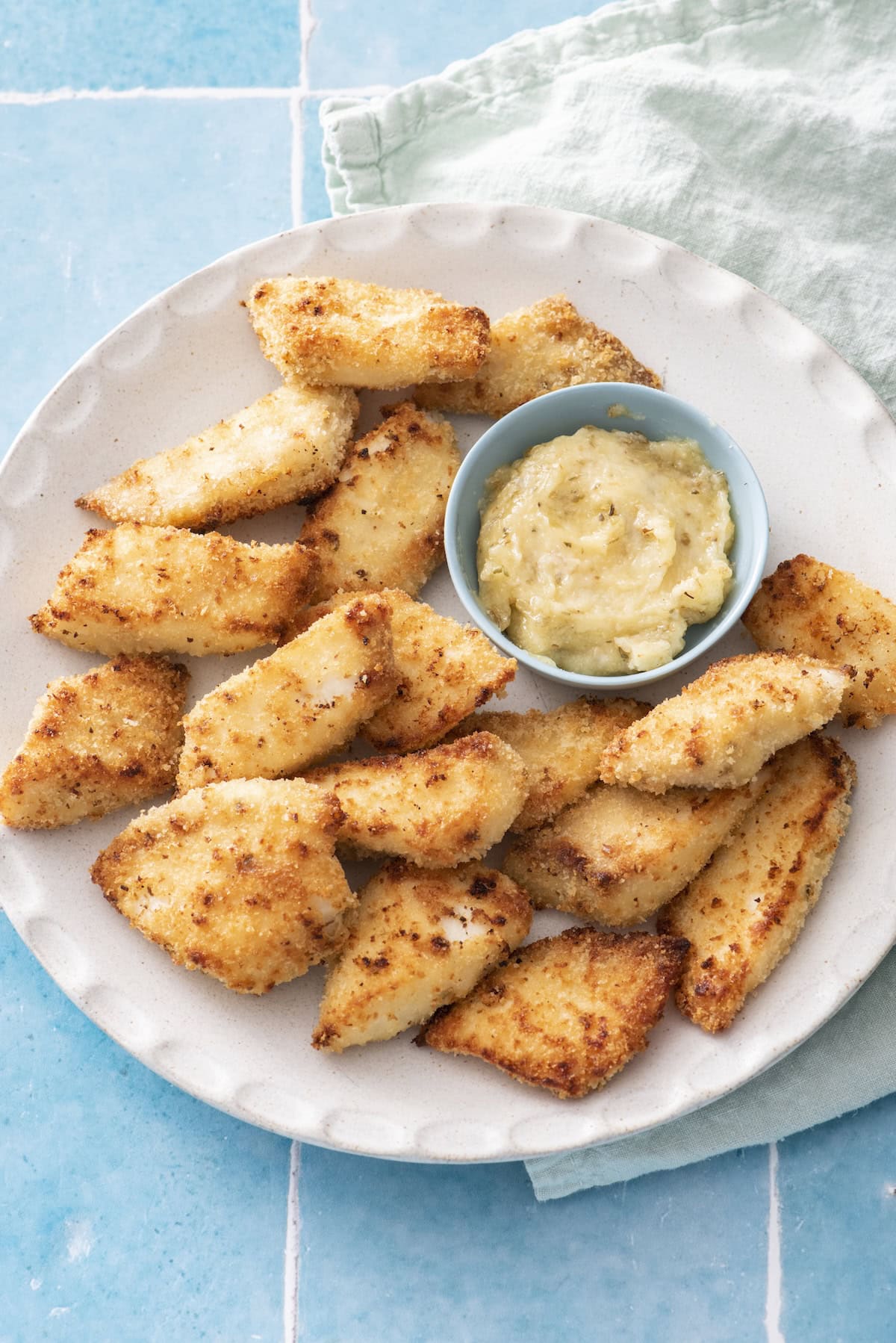 A plate of golden-brown breaded fish pieces with a small bowl of creamy dipping sauce, set on a light blue tiled surface with a pale green cloth napkin under the plate.