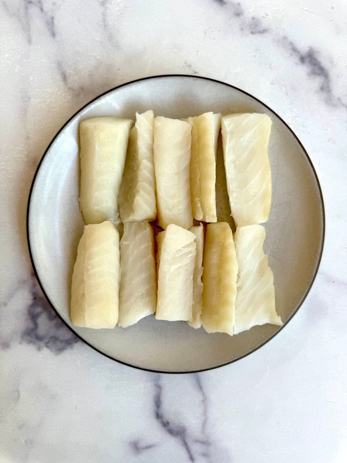 A plate with nine rectangular pieces of white fish, neatly arranged in three rows on a light-colored, marble-patterned surface.