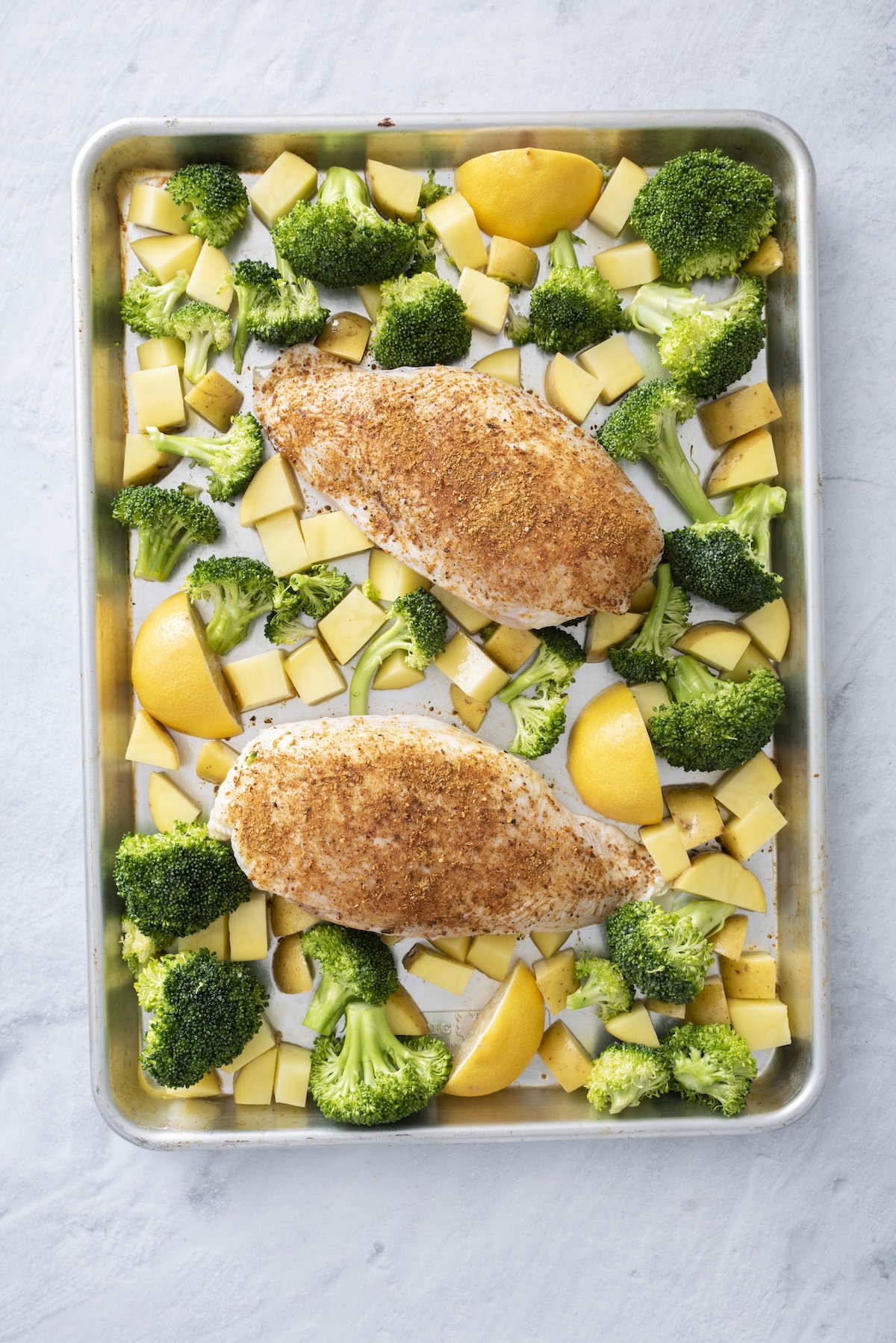 Raw za'atar-seasoned chicken breasts on a sheet pan with broccoli, potatoes, and lemon wedges before roasting.