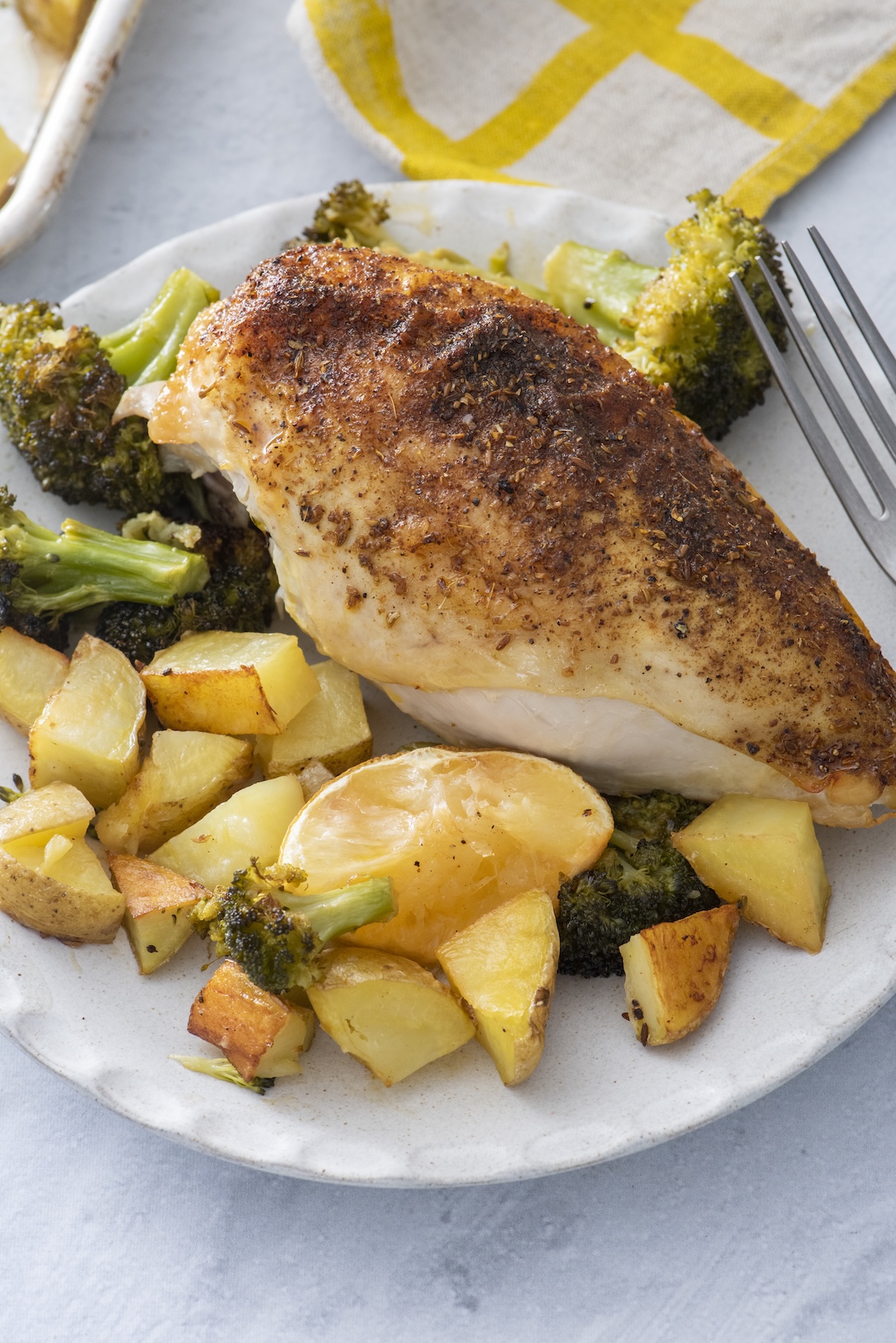 A za'atar roasted chicken breast served with crispy potatoes, broccoli, and a roasted lemon half on a plate.