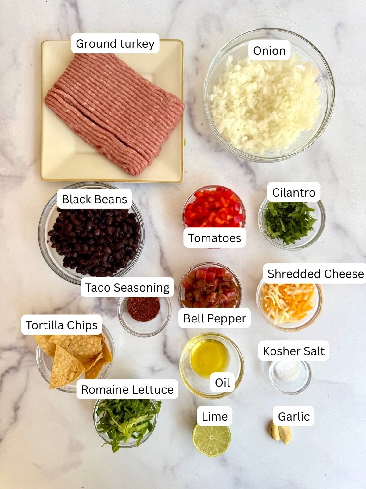 A flat lay of labeled taco bowl ingredients: ground turkey, diced onion, black beans, taco seasoning, tortilla chips, romaine lettuce, tomatoes, oil, bell pepper, lime, garlic, kosher salt, shredded cheese, and cilantro.
