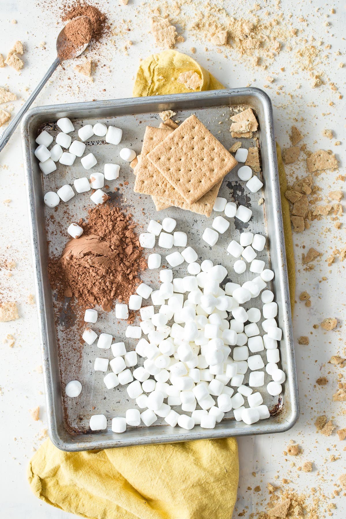 Graham crackers, marshmallows and cocoa powder displayed on baking sheet.