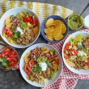 Three taco bowls with ground turkey, beans, tomatoes, chips, guacamole, and sour cream. Sides of chopped tomatoes, tortilla chips, guacamole, and sour cream on a patterned cloth.