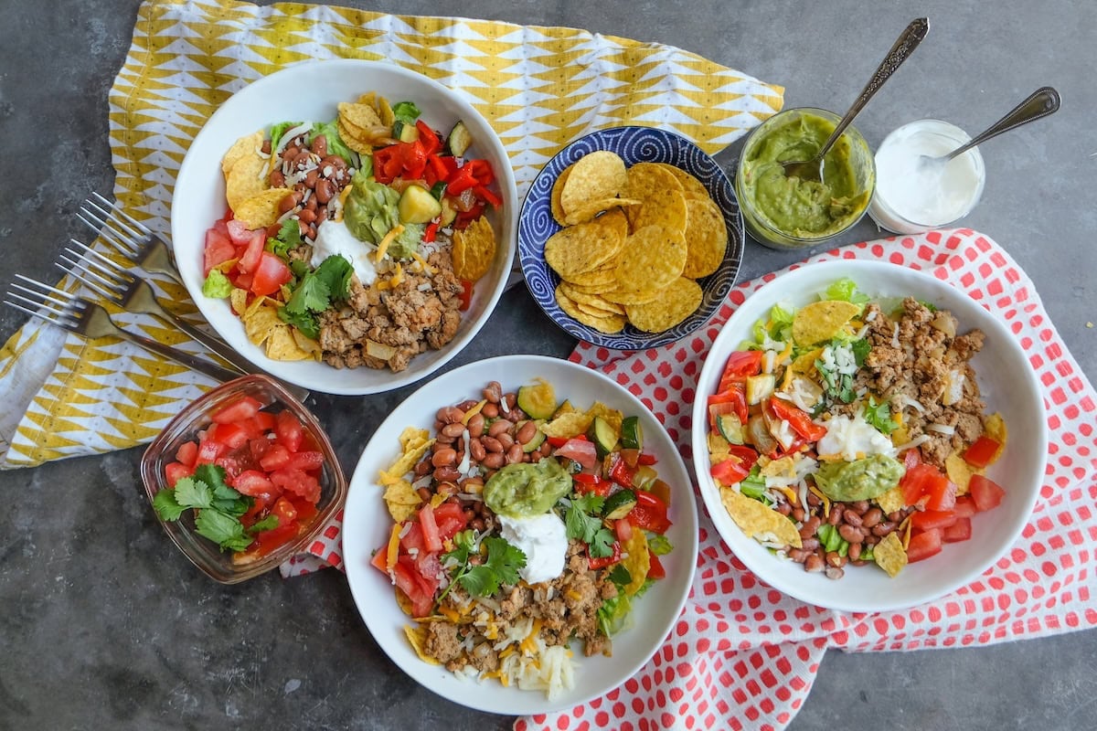 Three taco bowls with ground turkey, beans, tomatoes, chips, guacamole, and sour cream. Sides of chopped tomatoes, tortilla chips, guacamole, and sour cream on a patterned cloth.