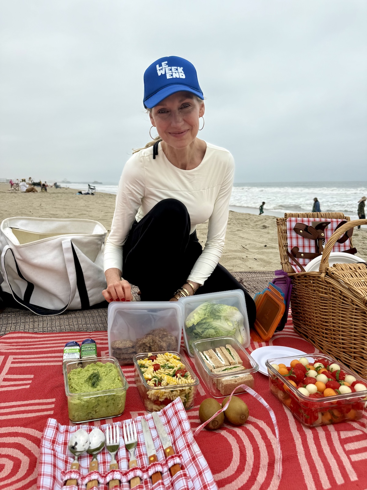 Catherine McCord sitting on a red picnic blanket at the beach with containers of pasta salad, cucumber sandwiches, fruit salad, and guacamole.