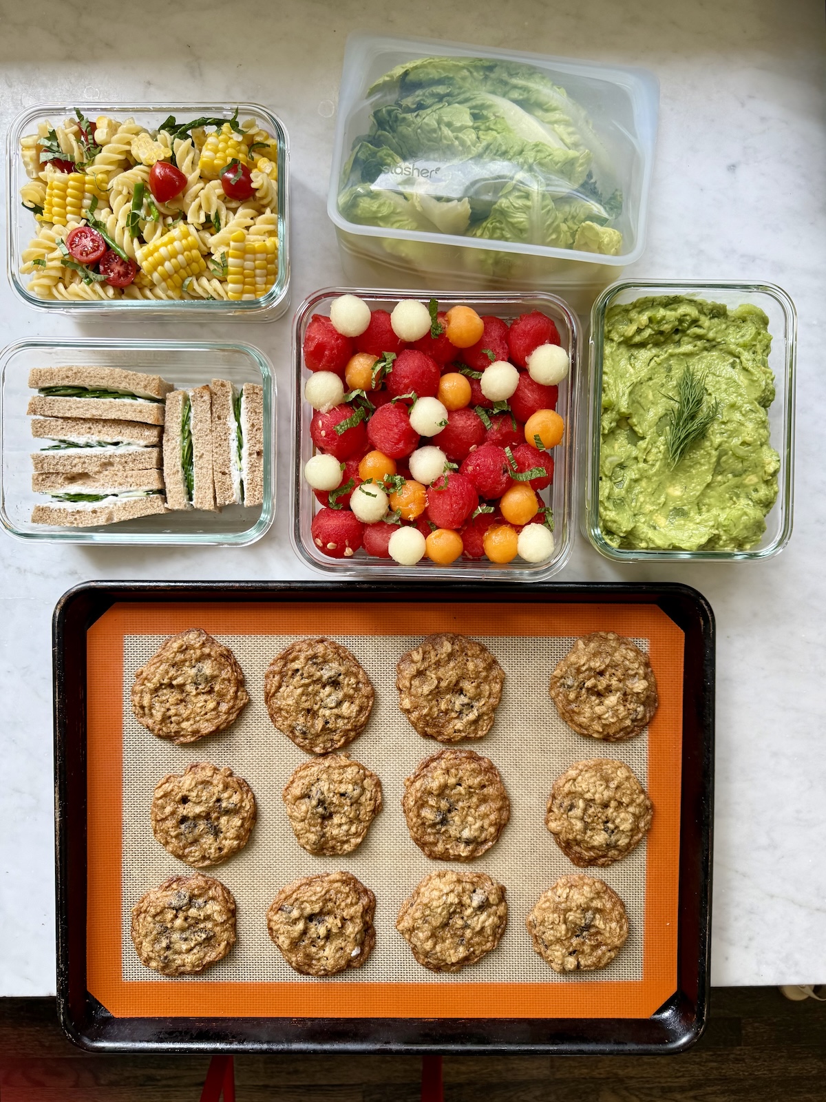 Overhead view of beach picnic foods including pasta salad, cucumber sandwiches, melon ball fruit salad, guacamole, and oatmeal cookies in glass containers.