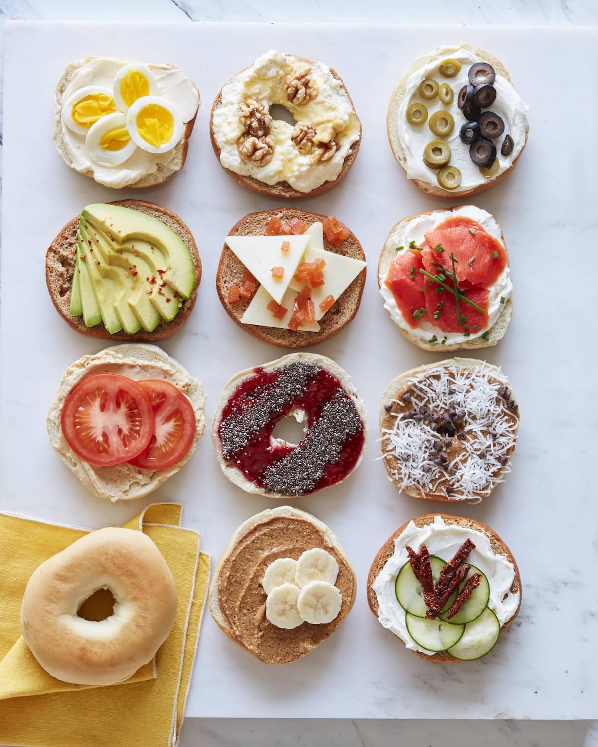 A dozen bagel halves topped with various ingredients, including eggs, walnuts, olives, avocado, cheese, smoked salmon, tomatoes, jam, banana, coconut, cucumber, and one plain bagel on a marble surface.