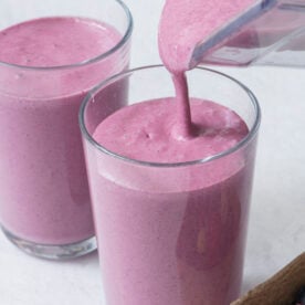 A thick purple smoothie is being poured from a blender into a glass, next to another full glass. A small container of fresh berries sits nearby.