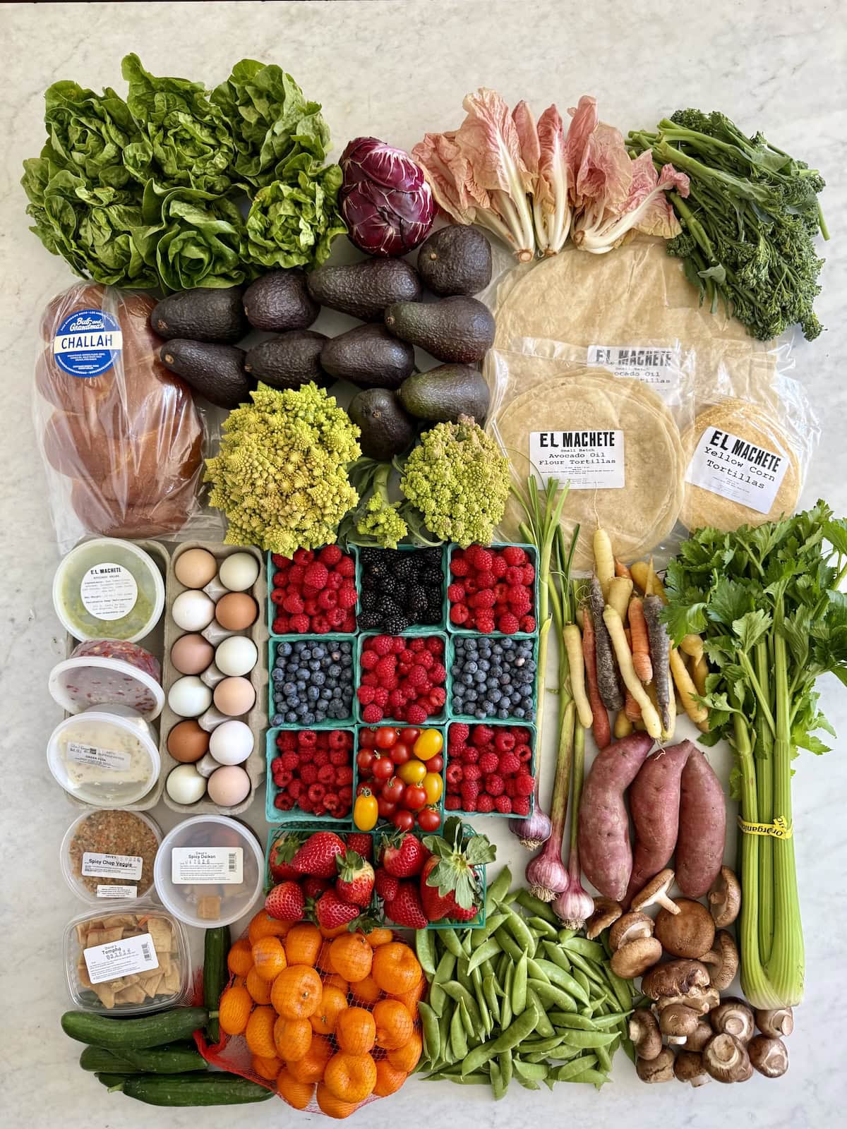 A farmers market haul of fresh produce laid out on a counter, including lettuce, avocados, berries, carrots, sweet potatoes, mushrooms, celery, citrus, eggs, and tortillas.
