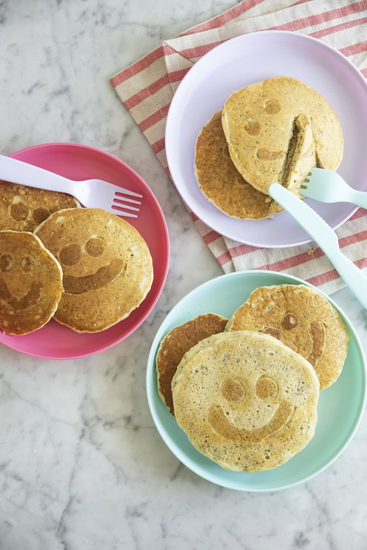 Three pastel plates with smiley face pancakes on a marble surface, one plate has a fork cutting a pancake, and a red-striped cloth is partly visible underneath a plate.