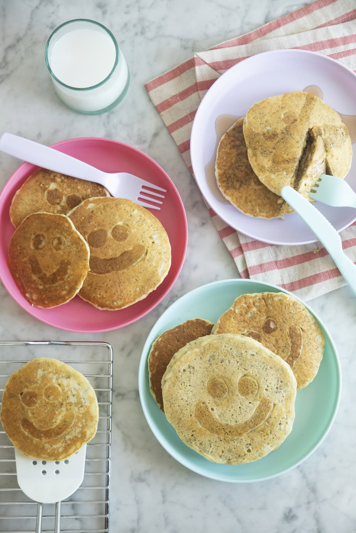 Three plates of smiley face pancakes with utensils, a cooling rack, and a glass of milk on a marble surface. One pancake is being sliced. Theres also a red-and-white striped napkin.