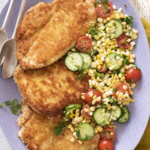 Three golden crispy chicken paillard cutlets on a platter with a fresh summer corn salad.
