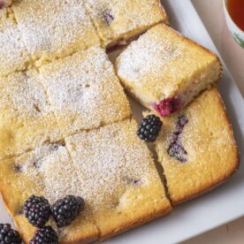 Blackberry tea cake cut into squares and dusted with powdered sugar on a white platter with fresh blackberries and a cup of tea.